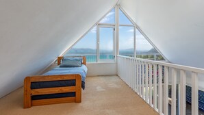 A bedroom with a sloped ceiling, a single bed, and a large window offering a view of the sea and distant mountains. - The Shack - at Seal Rocks (Seal Rocks)