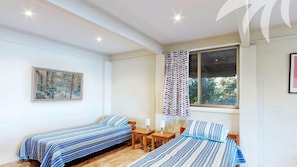 Quarto com duas camas de solteiro, um quadro na parede, uma janela com cortinas estampadas e uma pequena mesa com abajures.