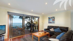 Sala de estar com uma grande porta de vidro deslizante que dá para uma varanda com vista para o mar. Há um sofá de couro preto, uma mesa de centro de madeira e uma televisão sobre um suporte.