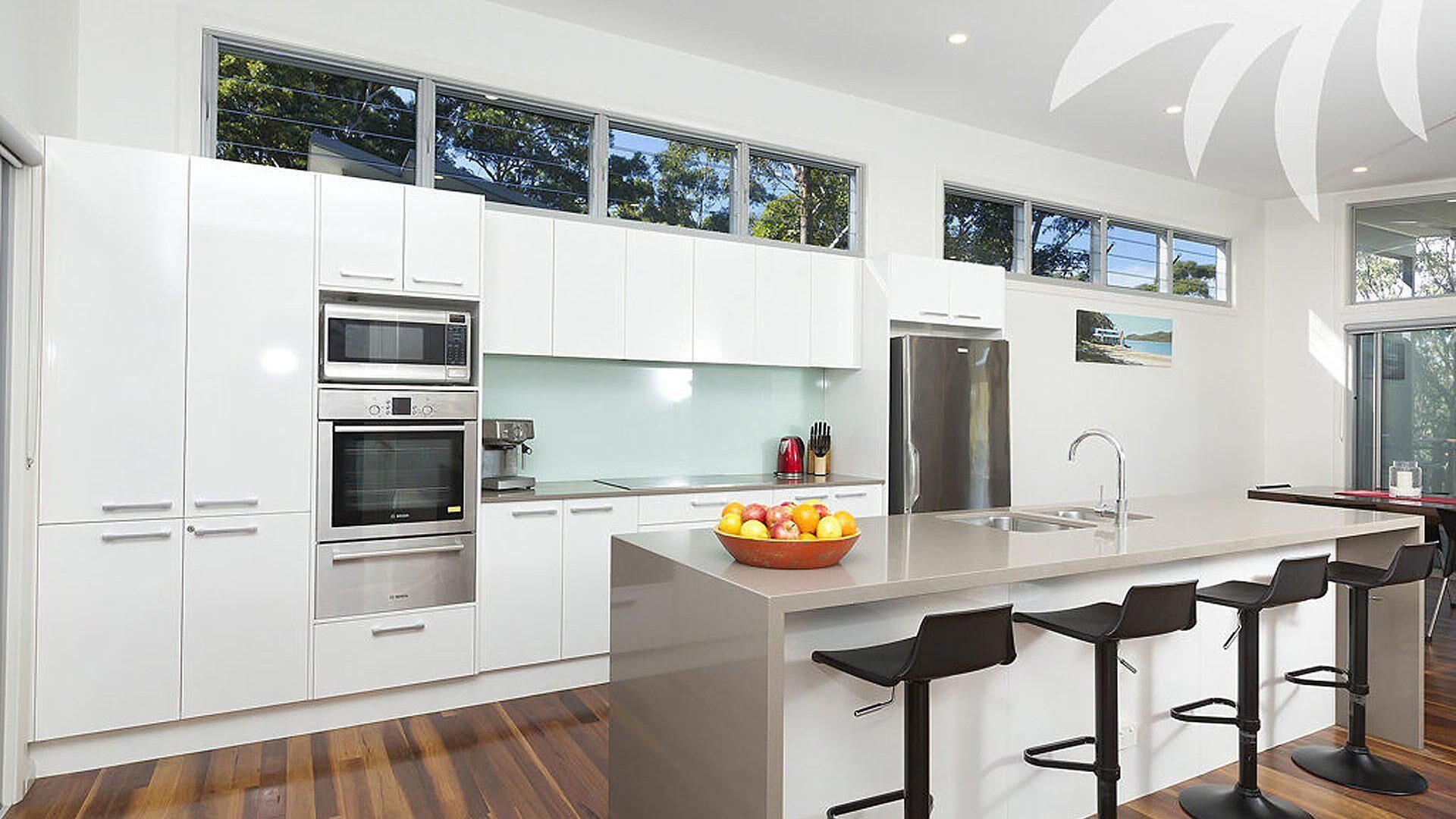 A modern kitchen with white cabinetry, stainless steel appliances, a large island with bar stools, and a view of the outdoors through large windows.