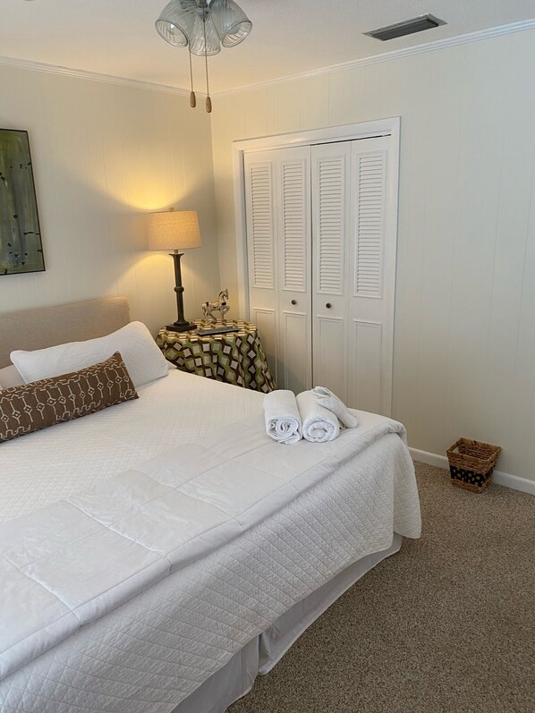 A neatly made bed with white bedding, a lamp, a small table with a patterned tablecloth, and a closed white door.