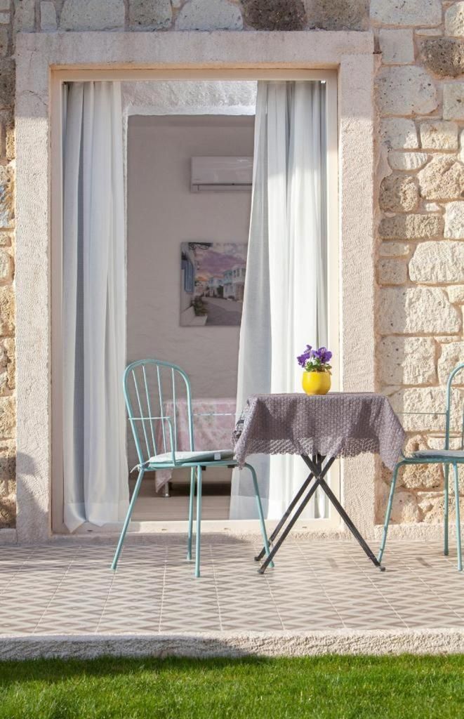 Un pequeño espacio al aire libre con mesa y sillas, una ventana con cortinas y una pared de piedra.