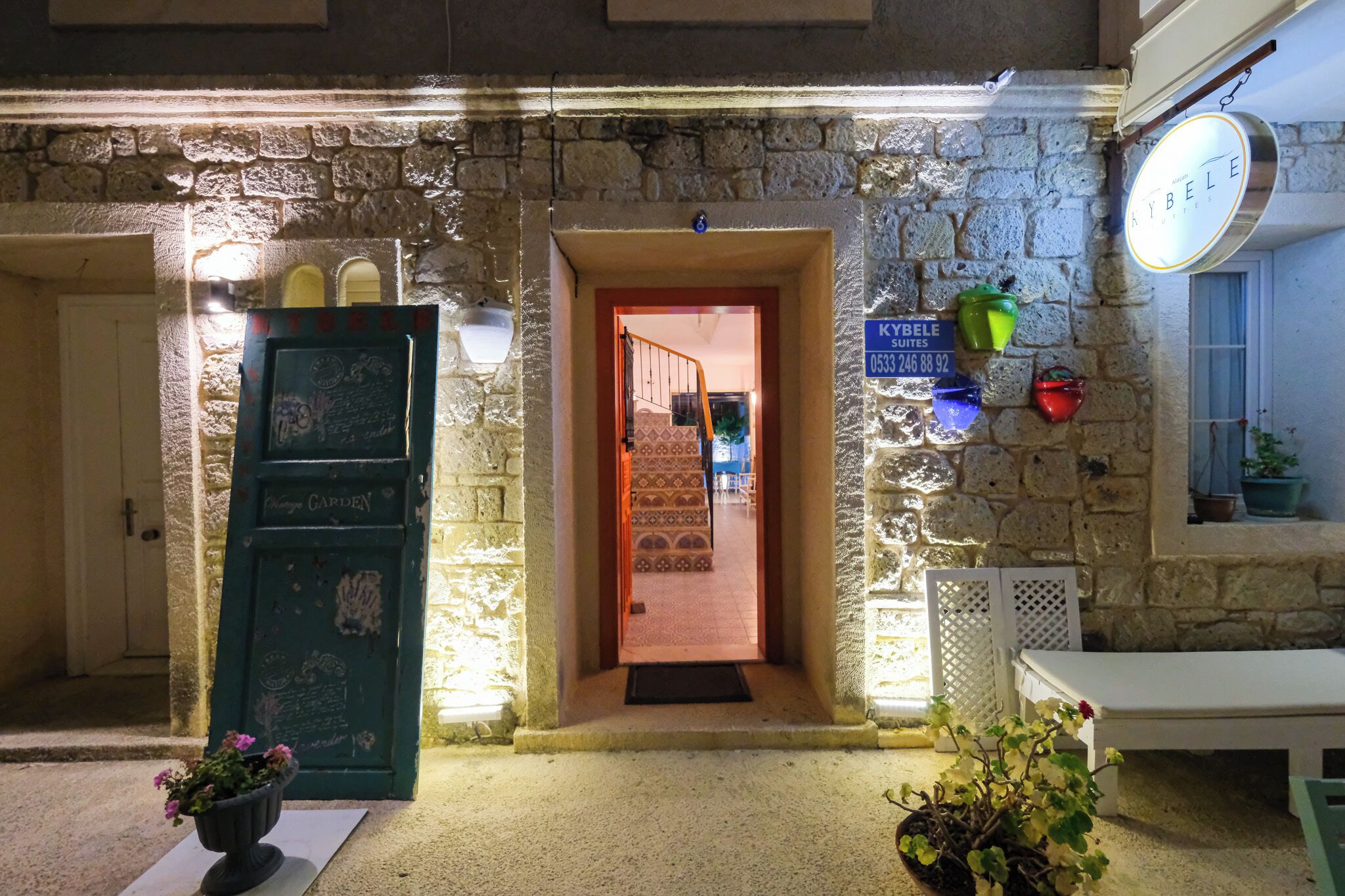 A stone building with a green door labeled "KYBELE SUITES," a sign with contact information, and a bench outside.