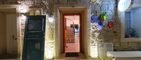A stone building with a green door labeled "KYBELE SUITES," a sign with contact information, and a bench outside.