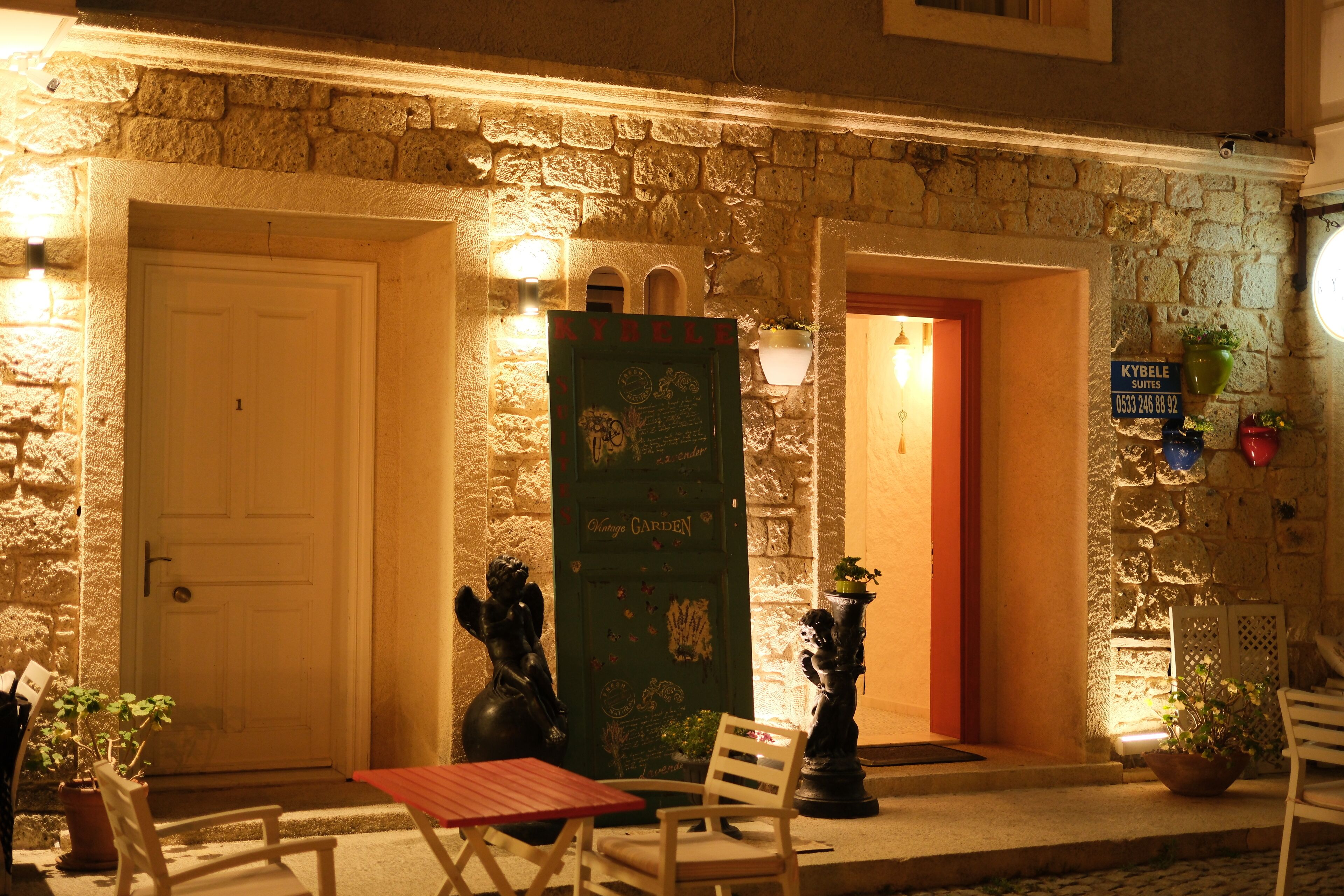 A stone building with a green door labeled "Kybale Suites" and a red door. There are potted plants and a bench outside.