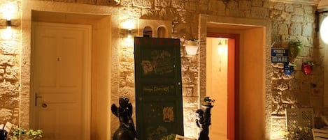 A stone building with a green door labeled "Kybale Suites" and a red door. There are potted plants and a bench outside.
