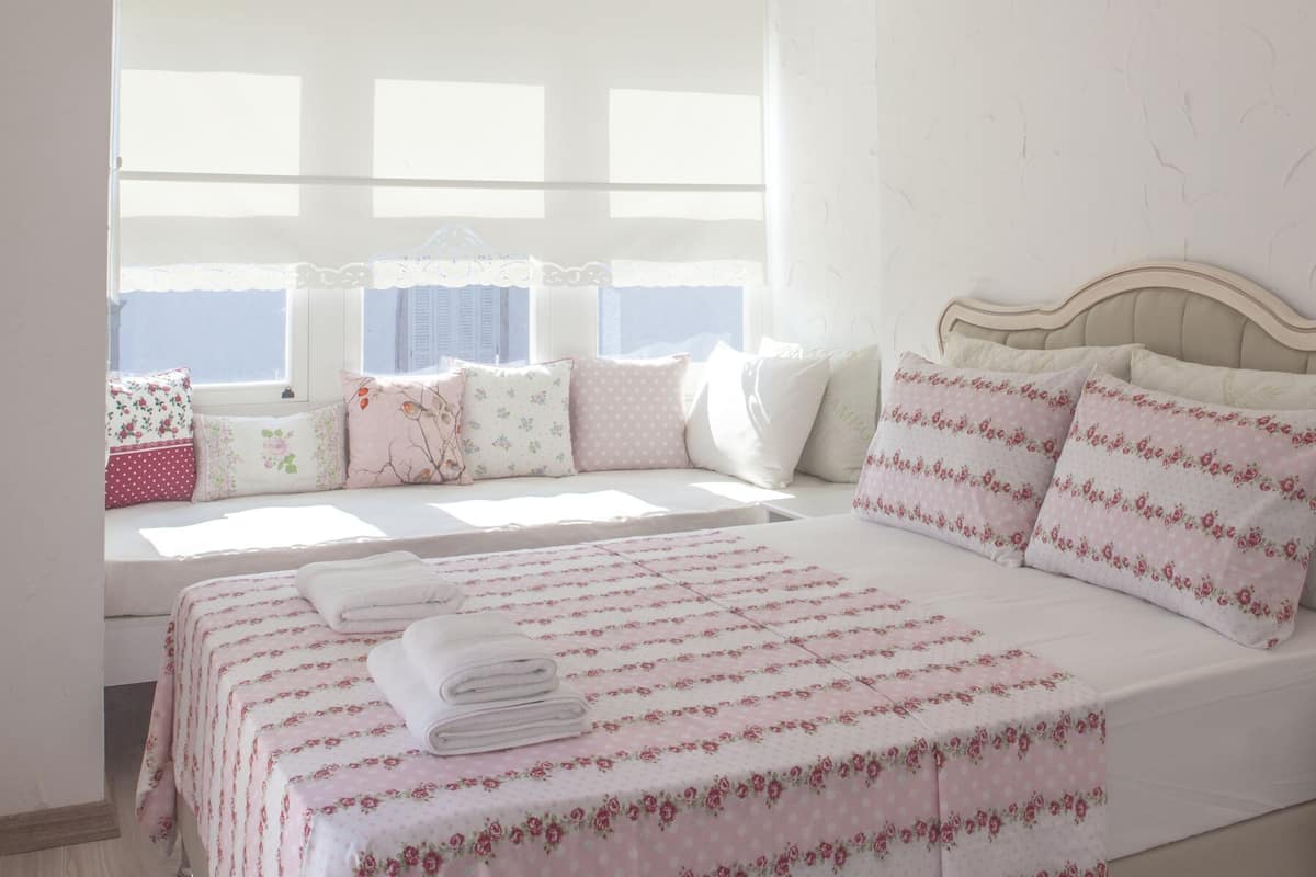 Een slaapkamer met een bed, kussens, een raam en een nachtkastje.