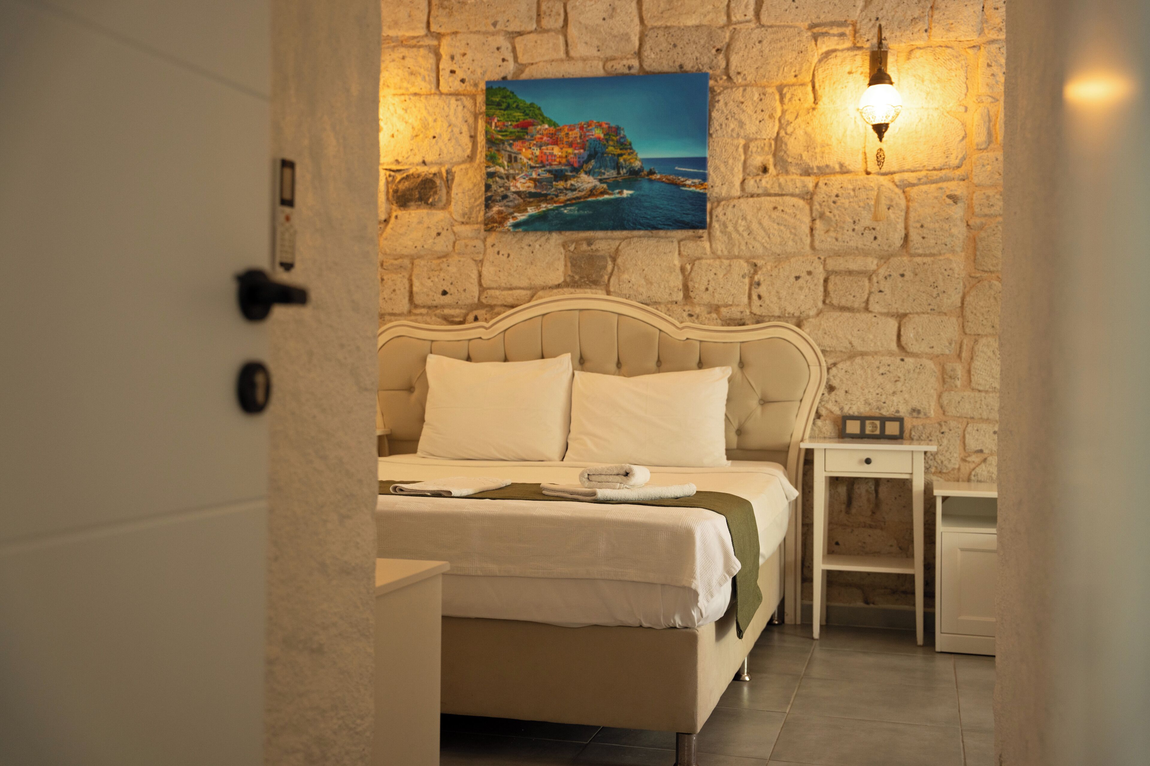 A hotel room with a stone wall, a bed with white linens, a painting of a coastal scene, and a nightstand with a lamp.