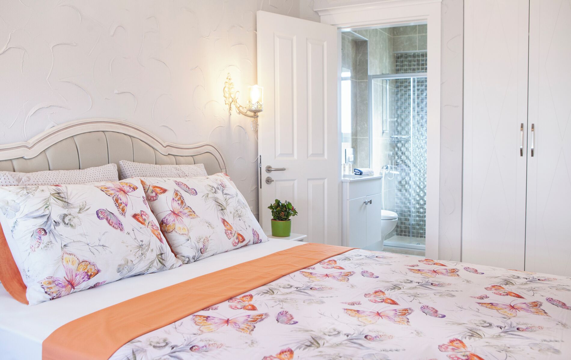 Ein Schlafzimmer mit Bett, Nachttisch, Topfpflanze und einem durchgegangenen Türrahmen sichtbarem Badezimmer.