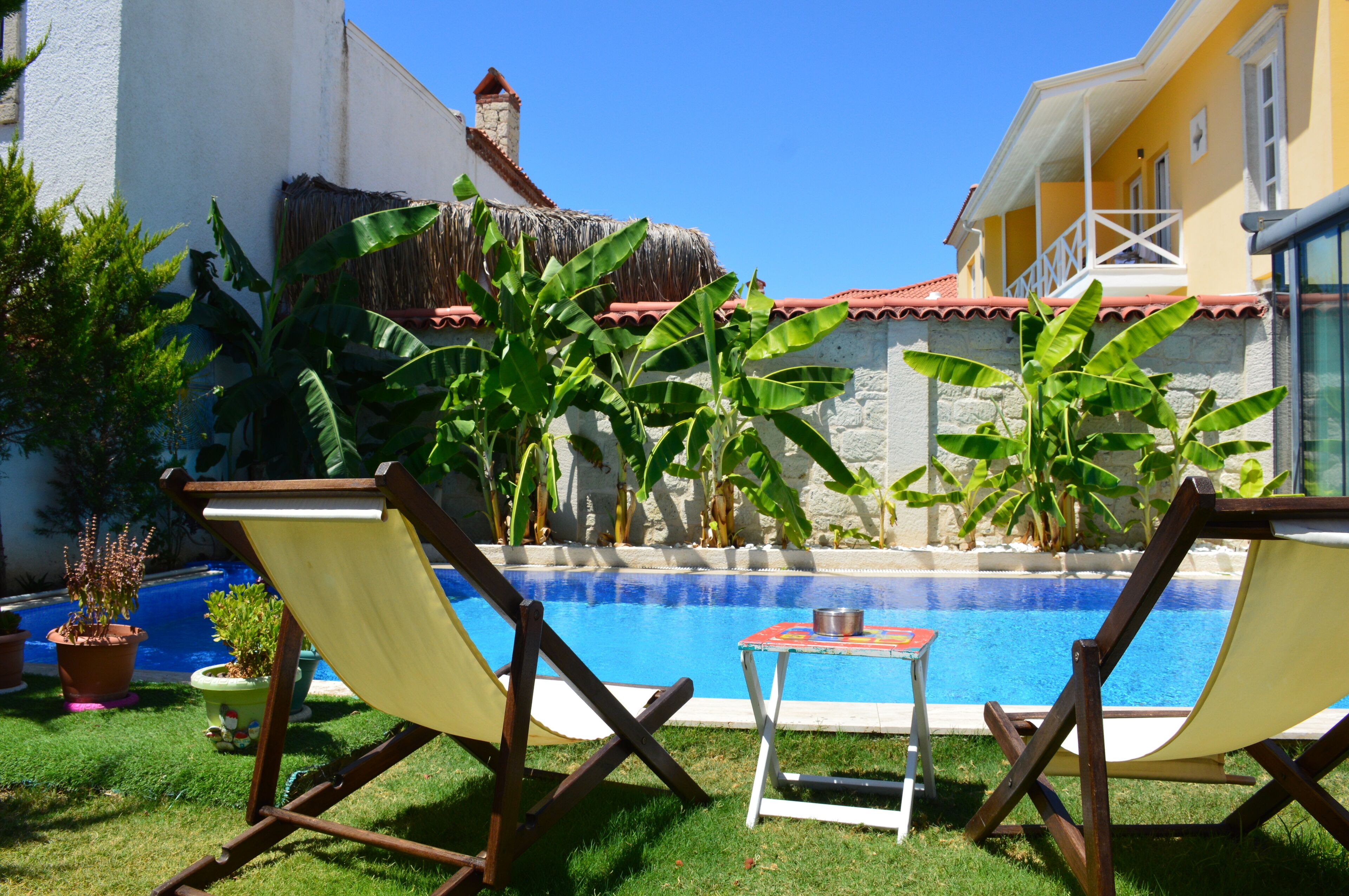 Un espace piscine comprenant deux chaises longues et une petite table, entouré de plantes en pot et d’un mur de pierre.