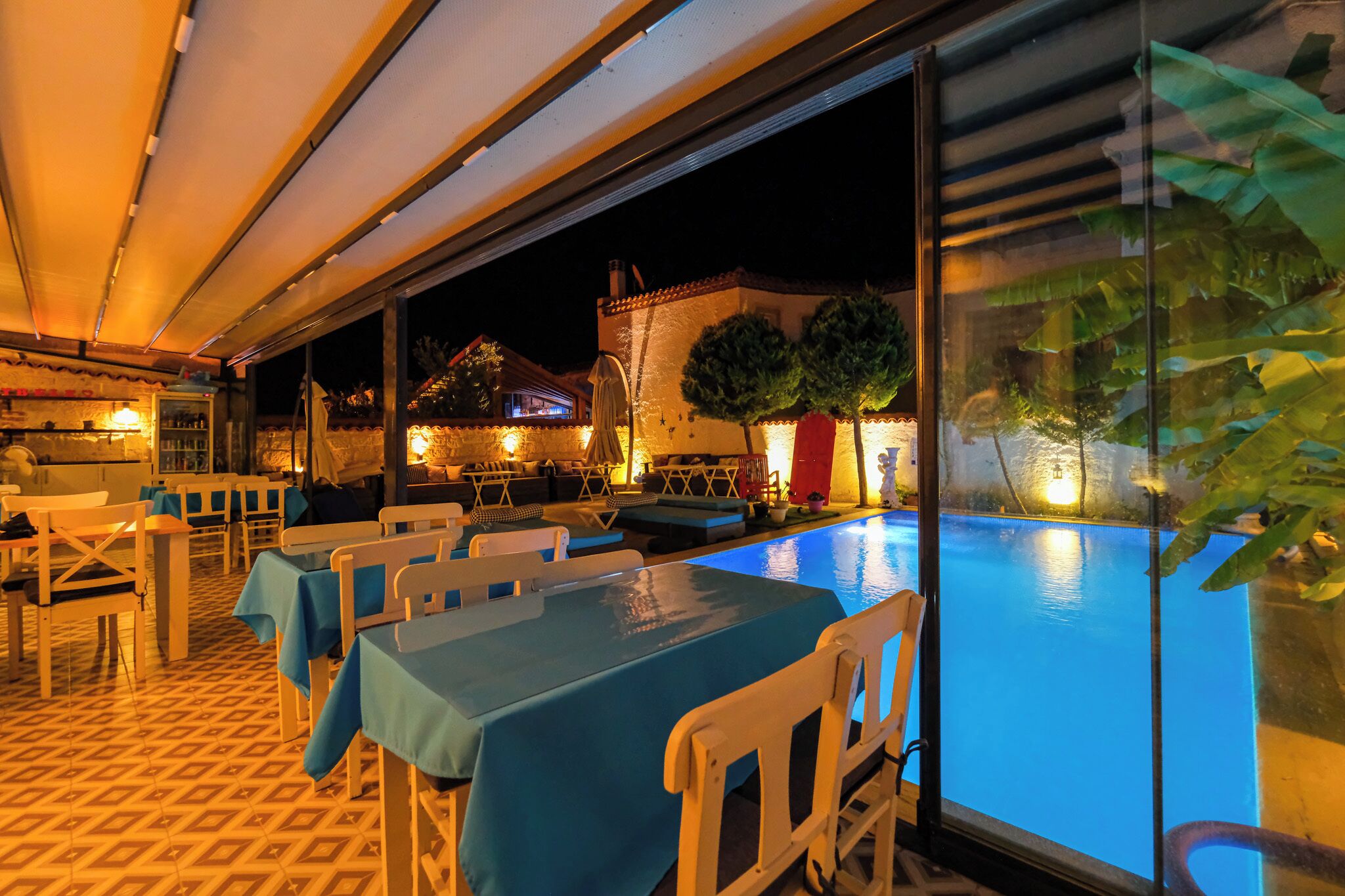 Un espace extérieur aménagé pour les repas, avec des tables et des chaises, une piscine et une vue sur un bâtiment illuminé la nuit.