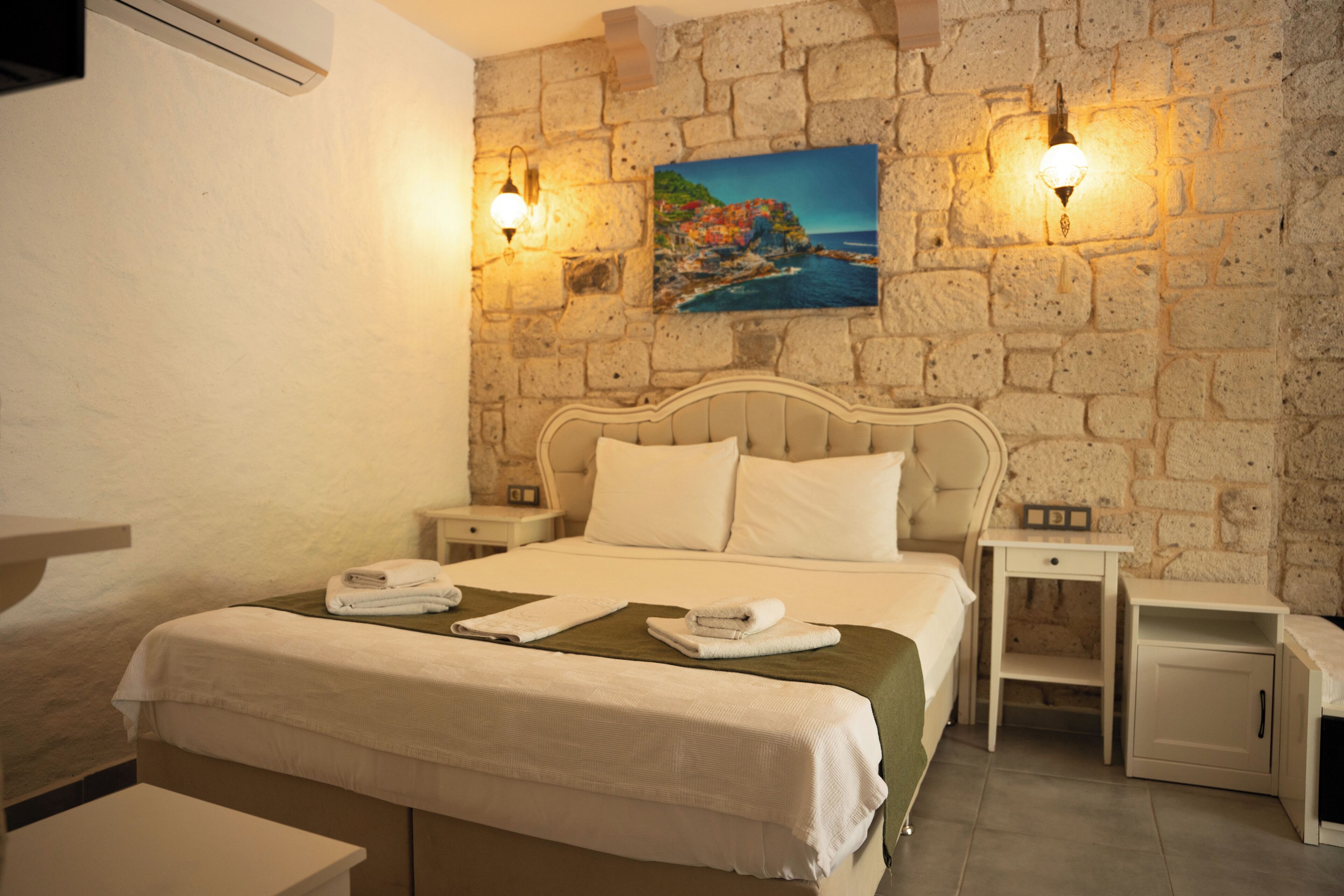 Habitación de hotel con pared de piedra, cama con ropa blanca, dos mesitas de noche y un cuadro de una escena costera.