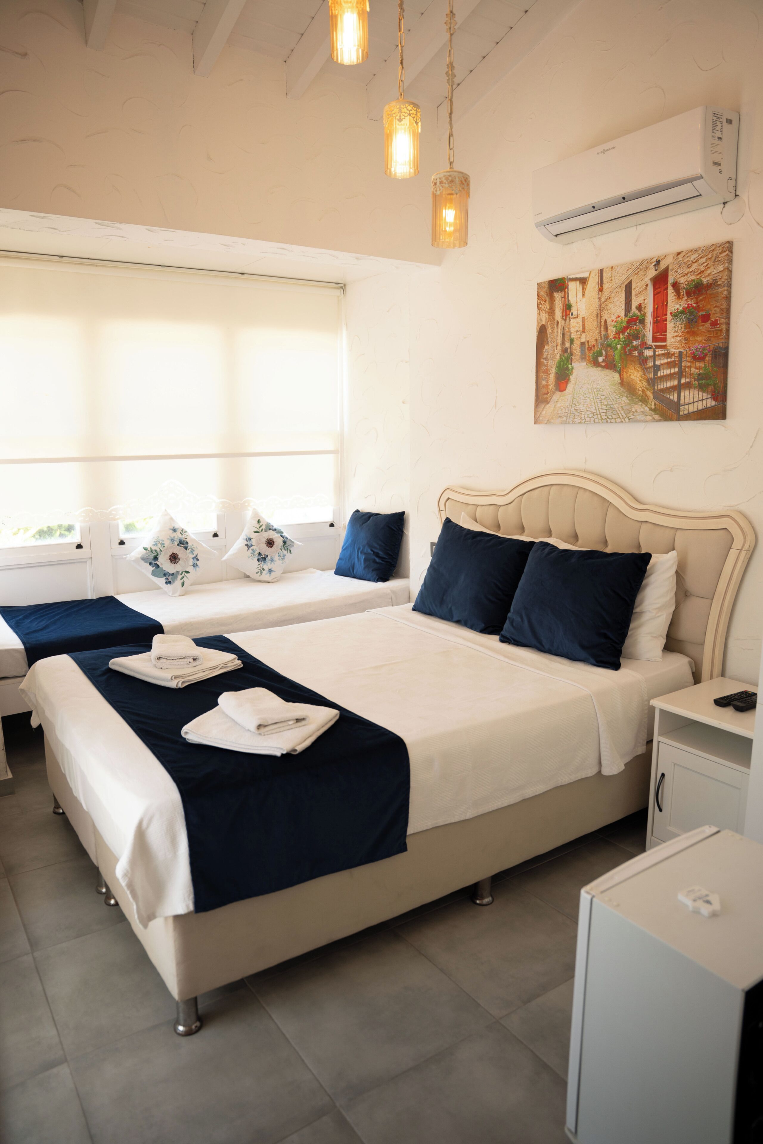 Une chambre d’hôtel avec deux lits, une fenêtre avec des stores, un tableau au mur et un appareil de climatisation.