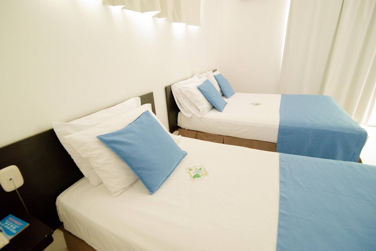 A hotel room with two single beds, each with a blue and white bedspread, and a nightstand with a phone and a book.