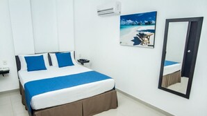 A neatly arranged hotel room with a bed, a nightstand, a wall-mounted air conditioner, a framed picture of a beach, and a mirror. - Hotel Maranatá (Valledupar)