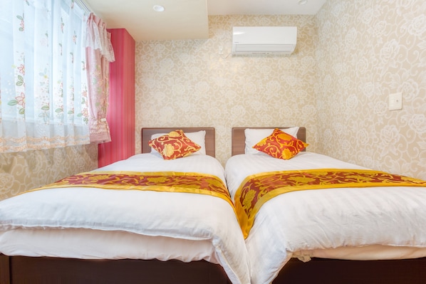 A hotel room with two beds, each with white bedding and a red and yellow patterned blanket. There is a window with a floral curtain, and an air conditioning unit mounted on the wall.