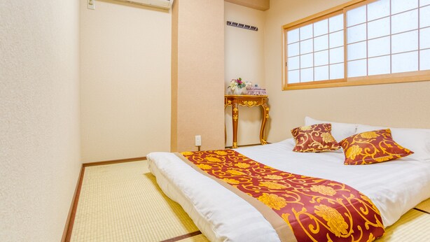 Una habitación tradicional japonesa con suelo de tatami, una cama con colcha de motivos rojos y dorados, y una ventana con diseño de cuadrícula.