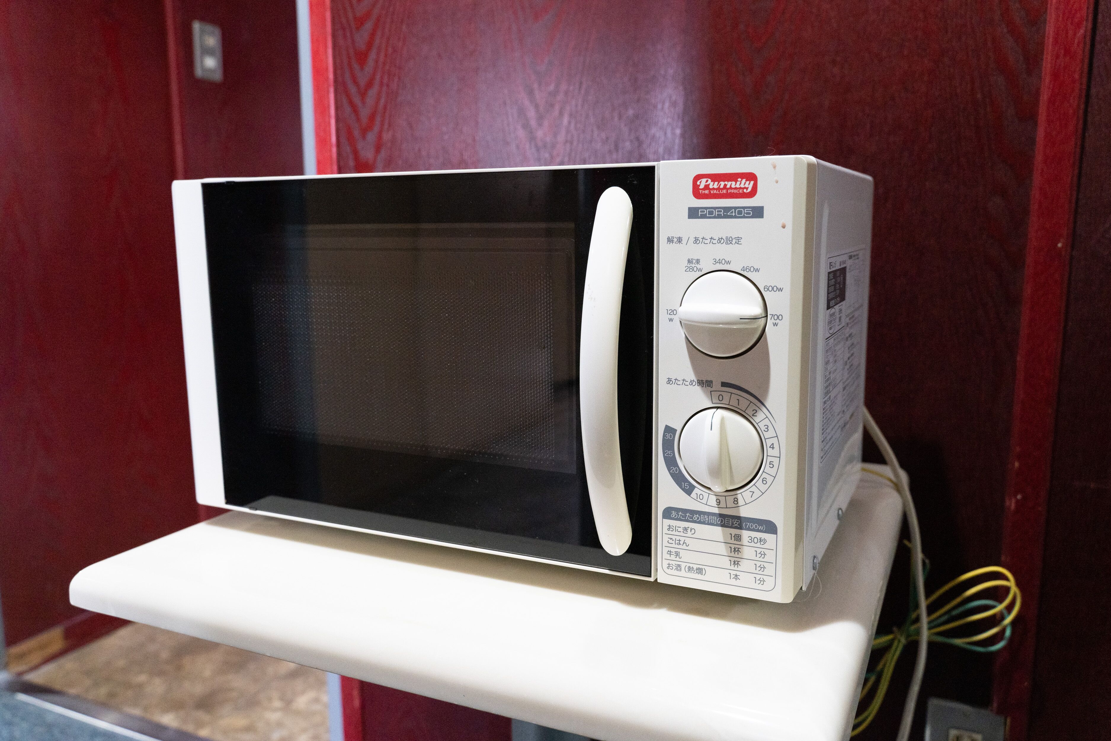 A white microwave oven with a digital display and control panel, mounted on a white shelf against a red wall.