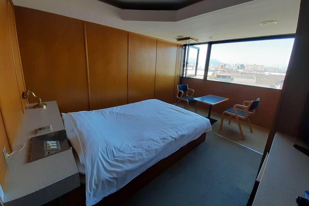 A hotel room with a bed, a desk, a chair, a small table, and a view of the city through large windows.