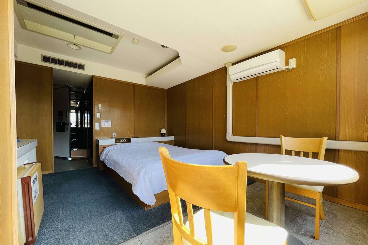 A compact hotel room with a single bed, a small table with a chair, and a built-in air conditioning unit.