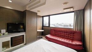 A hotel room with a bed, a red sofa, a television, and a city view. - Hotel Sun (Takasaki)