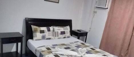 A hotel room with a bed, a nightstand, an air conditioning unit, and a curtain.