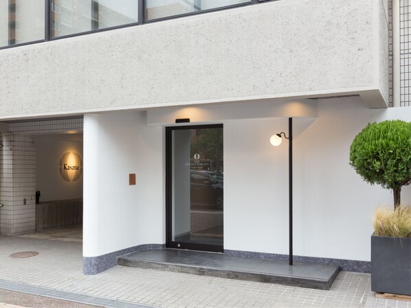 A modern hotel entrance with a glass door, a sign for 'The Hotels Hakata', and a small tree planter on the right. - The Hotels Hakata Kasane Bayside (Fukuoka)