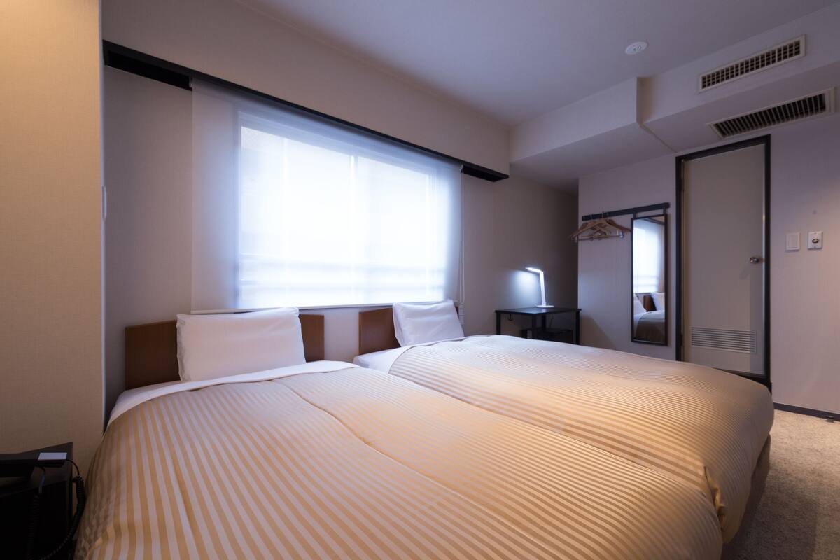 A hotel room with two beds, a bedside table with a lamp, a mirror, and a wardrobe.