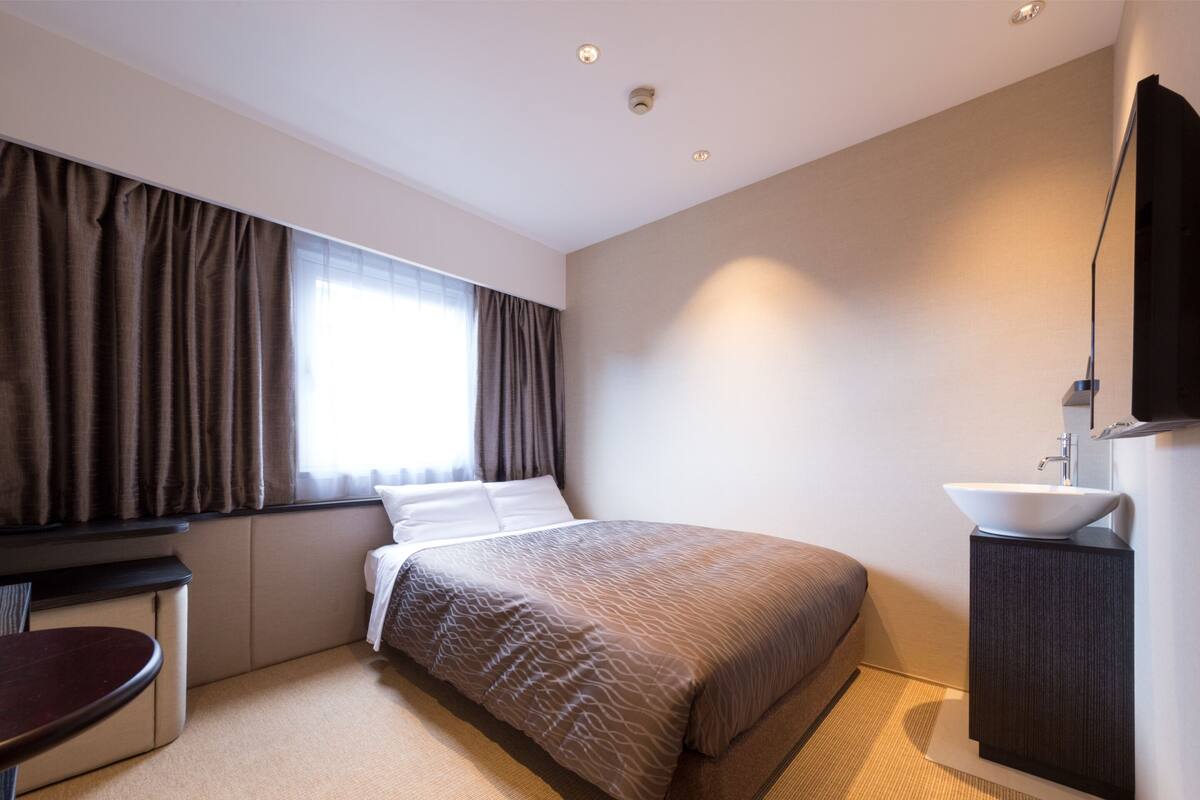 A modern hotel room with a large bed, a bedside table, a small desk, a bathroom with a sink, and a large window with curtains.