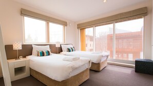 Habitación de hotel con dos camas, una mesita de noche, una lámpara y una ventana con vistas a edificios.