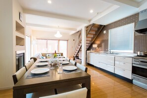 A modern kitchen with a wooden dining table set for four, a staircase, and a large window with blinds. - Snow Fox (Kutchan)