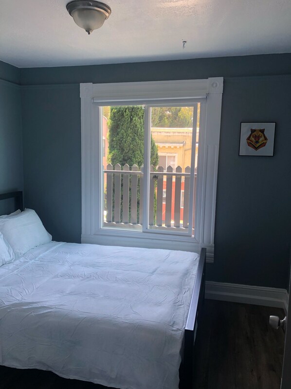 A bedroom with a bed, a window with a view of a fence and trees, and a wall-mounted picture.