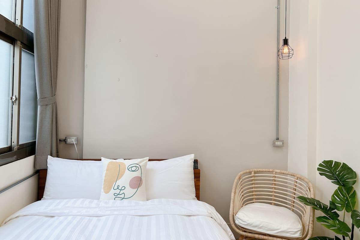 Quarto com cama, cadeira, janela com cortinas e luminária fixada na parede.