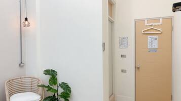 Una habitación con puerta de madera, perchero colgante, planta en maceta, silla de mimbre y aplique de pared.