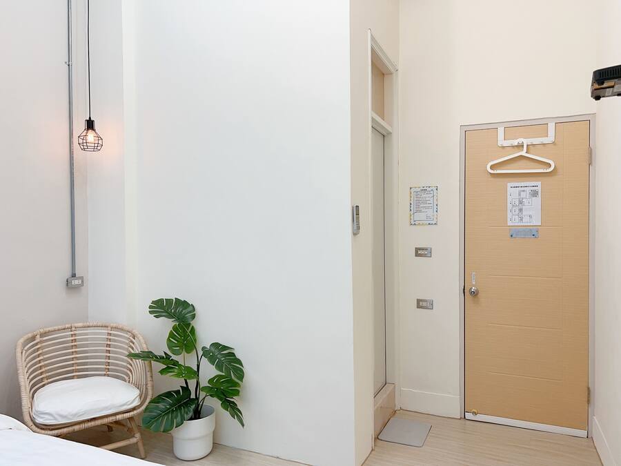 Una habitación con puerta de madera, perchero colgante, planta en maceta, silla de mimbre y aplique de pared.