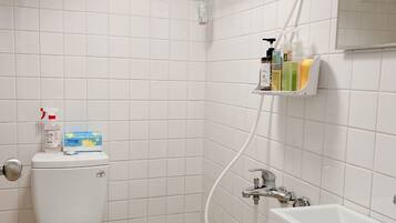 Un baño pequeño y limpio con inodoro, lavamanos y ducha. Hay artículos de aseo en la pared y un perchero para toallas.