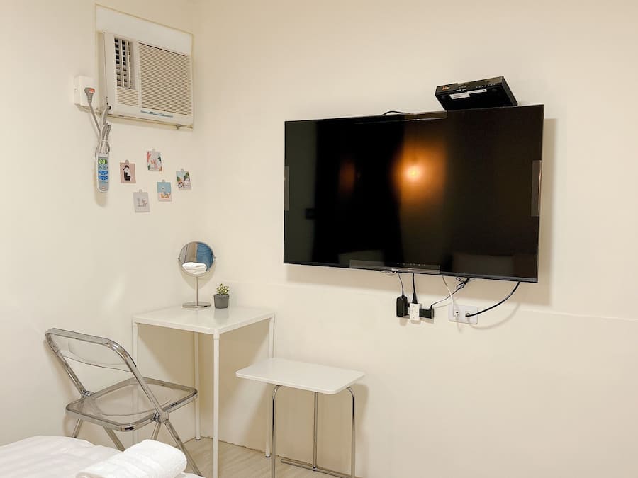 A hotel room with a bed, a TV mounted on the wall, a small desk, a chair, and a wall-mounted air conditioning unit.