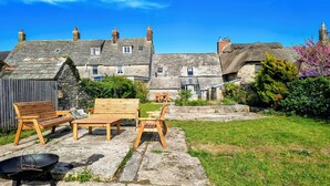 Une maison en pierre avec un toit de chaume, un jardin agrémenté de meubles en bois et un espace barbecue.