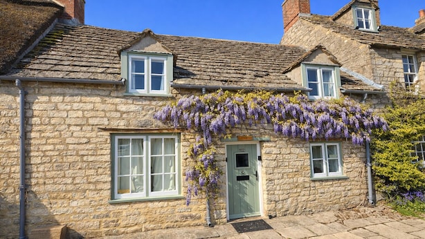 Un bâtiment en pierre avec une porte verte, deux fenêtres et un petit porche orné de fleurs violettes.