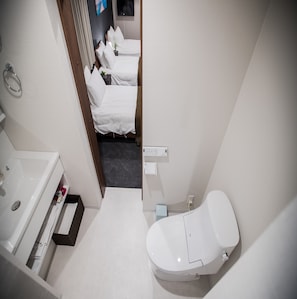 A modern bathroom with a white sink, toilet, and shower area. There is a small bedside table with a plant and a mirror above the sink.