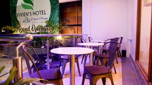 Un hall de hotel con un cartel que dice 'Vivien's Hotel' y 'Gran Inauguración'. Hay mesas y sillas redondas dispuestas para los huéspedes.