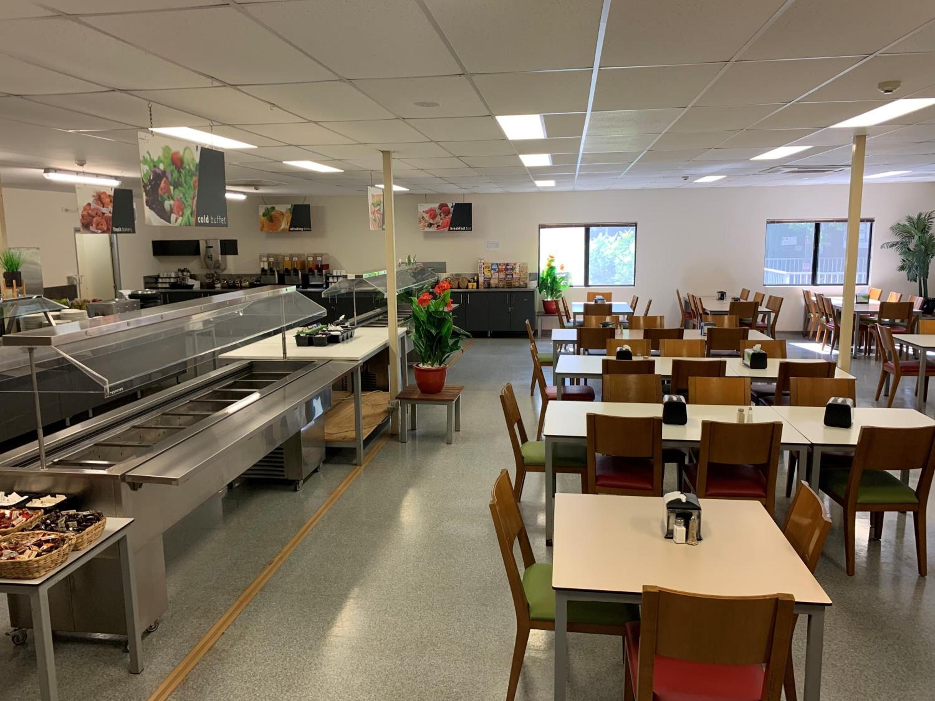 A cafeteria with stainless steel serving lines, tables, chairs, and potted plants.