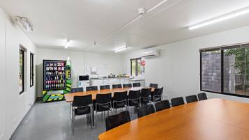 Un salón de conferencias con una larga mesa de madera, sillas negras, una máquina expendedora y una cocina pequeña al fondo.