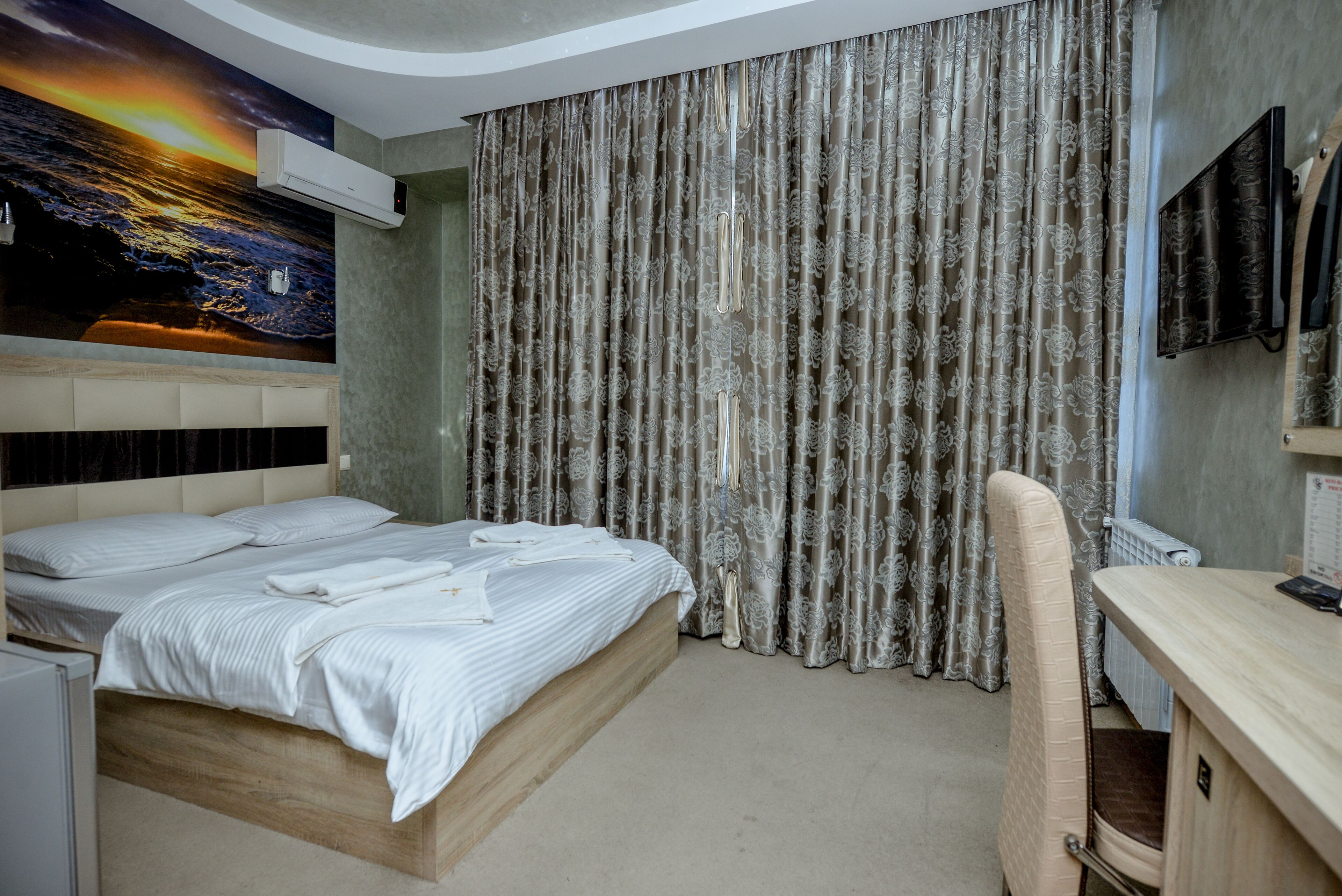 Quarto de hotel com cama, escrivaninha, cadeira, televisão e cortina estampada com flores.
