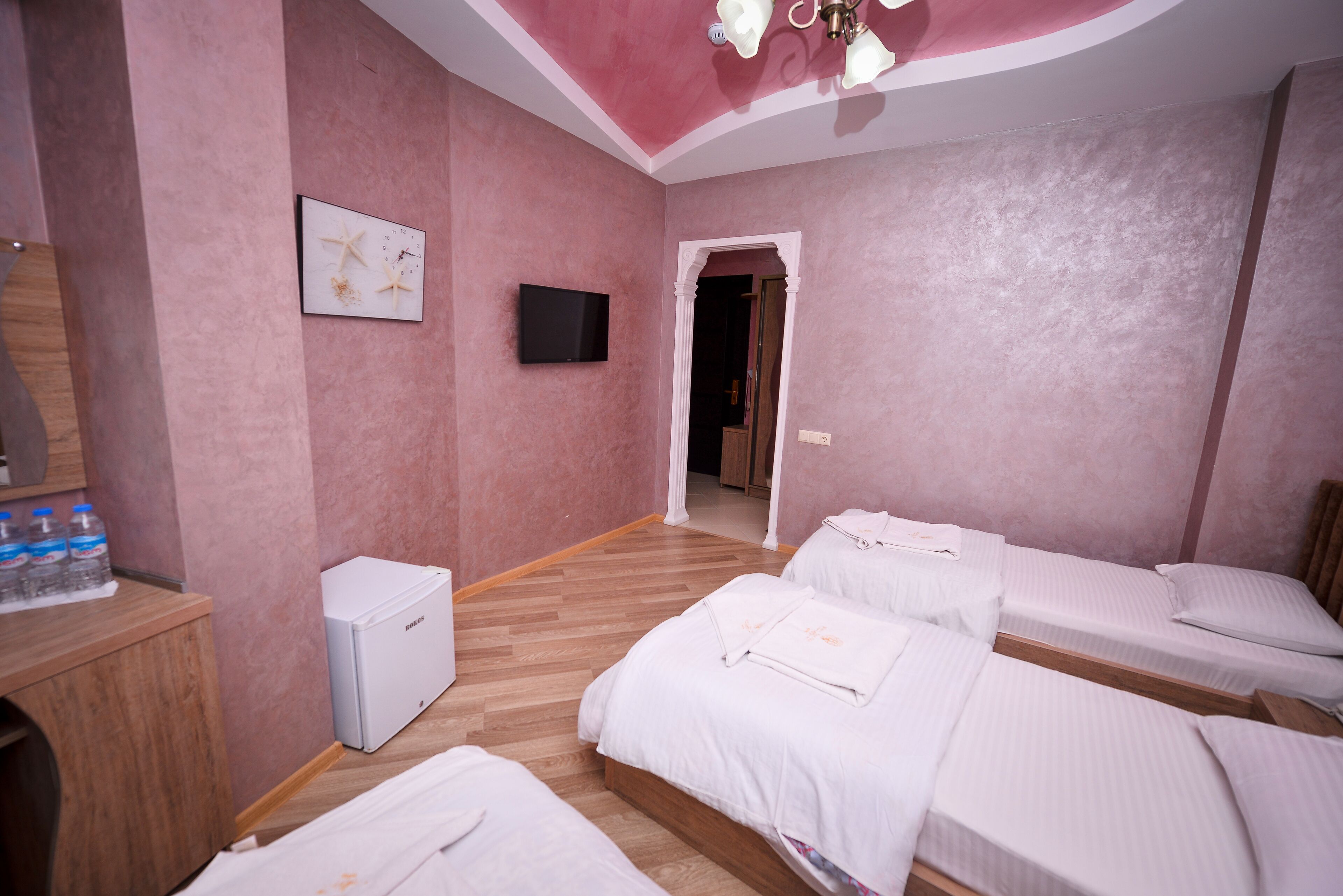 A hotel room with pink walls, a wooden floor, a TV mounted on the wall, and three beds.