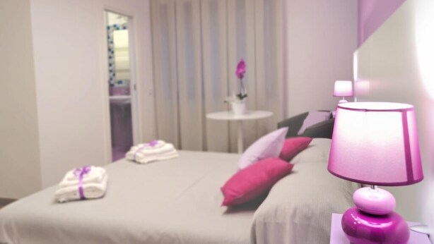 Camera d'albergo con un letto, cuscini, un comodino con una lampada e un piccolo tavolo con un vaso.