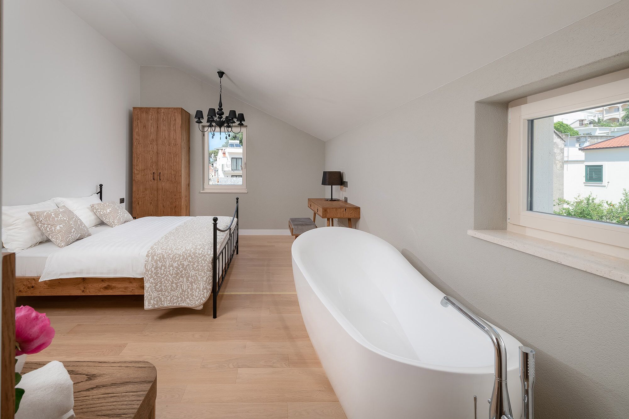 A bedroom with a large bed, a wooden wardrobe, a window with a view, a bedside table with a lamp, and a freestanding bathtub.