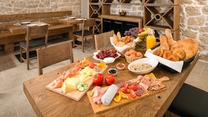 Una mesa de madera con una variedad de alimentos, como quesos, carnes y panes, frente a una pared de piedra con una estantería para vinos.