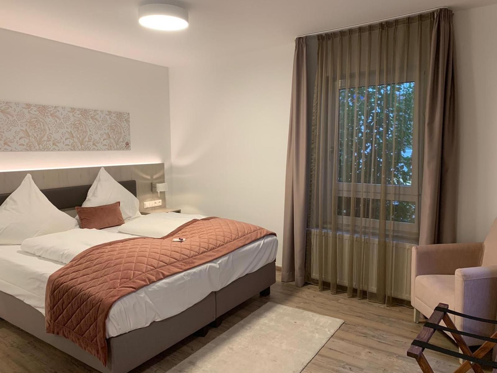 Ein modernes Schlafzimmer mit Bett, Nachttisch, Lampe und Blick auf Bäume durch ein Fenster.