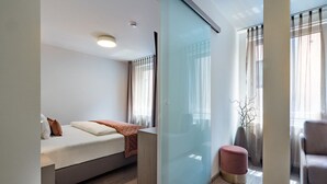 A modern bedroom with a bed, a glass partition, and a seating area with a chair and ottoman. - Boutique Hotel Rose Baden-Baden (Baden-Baden)
